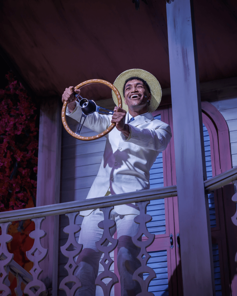 Once on This Island at The Ruth: Storytelling as Survival Through Music and Memory 4 A performer in a white suit and straw hat joyfully holds a steering wheel while standing on a balcony, with a floral backdrop.