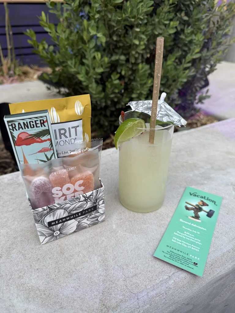 Meanwhile Park and the Art of Gathering: A Conversation with Jeff Paris and Matthew Ivan Bennett 2 A refreshing drink in a glass with a straw and a paper umbrella, accompanied by colorful candy packets and a green promotional card, placed on a light-colored surface with greenery in the background.