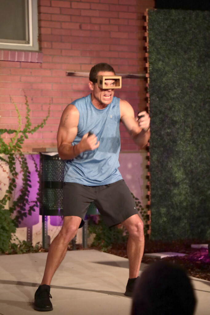 Meanwhile Park and the Art of Gathering: A Conversation with Jeff Paris and Matthew Ivan Bennett 6 A man wearing a blue sleeveless shirt and shorts is performing an energetic workout outside, with a wooden frame over his eyes that appears to simulate binocular vision.