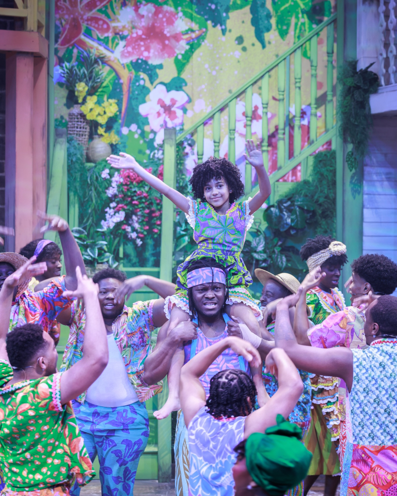 Once on This Island at The Ruth: Storytelling as Survival Through Music and Memory 7 A vibrant stage performance featuring a group of dancers and singers in colorful tropical outfits. A man carries a smiling girl on his shoulders while others engage joyfully around them, set against a floral backdrop.