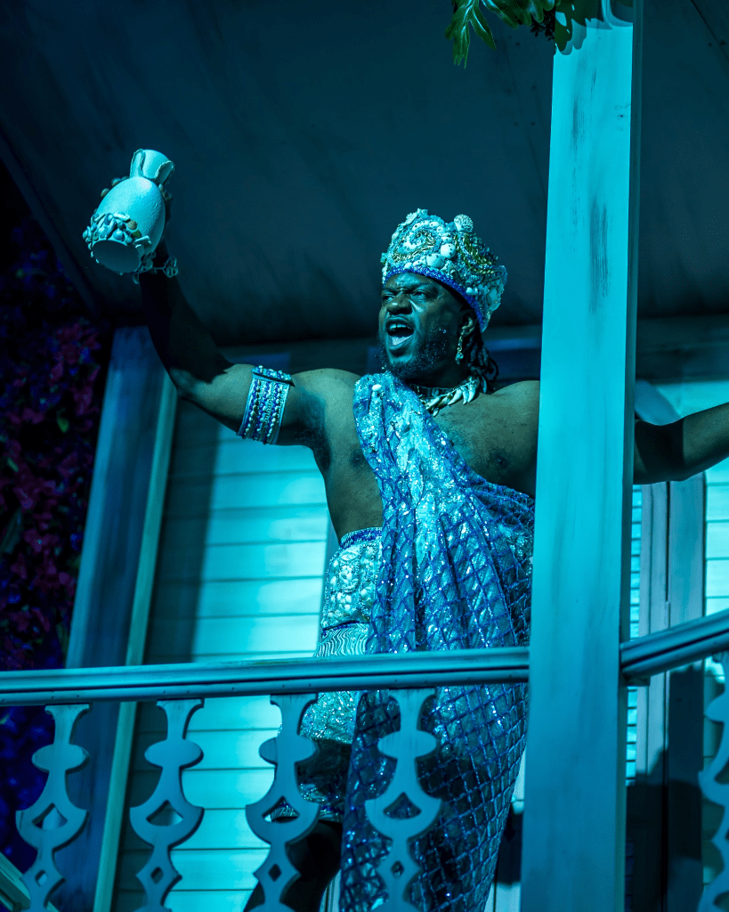 Once on This Island at The Ruth: Storytelling as Survival Through Music and Memory 6 A performer dressed in a shimmering blue outfit and crown stands on a balcony, holding a pottery jar and celebrating.
