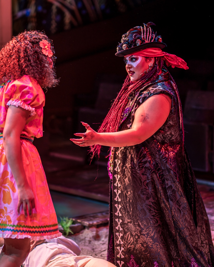 Once on This Island at The Ruth: Storytelling as Survival Through Music and Memory 5 Two performers on stage in a theatrical production, one in a colorful dress with a floral accessory and the other in a dramatic costume with a black cape and elaborate hat, engaged in an intense conversation.
