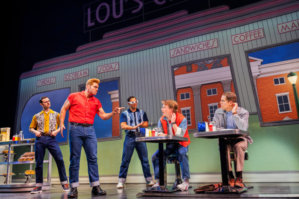 A scene from a musical featuring five male characters in casual 1950s attire, performing in front of a diner backdrop labeled 'Lou's'. Two characters are seated at tables, while three others are standing, engaging in dynamic interaction.