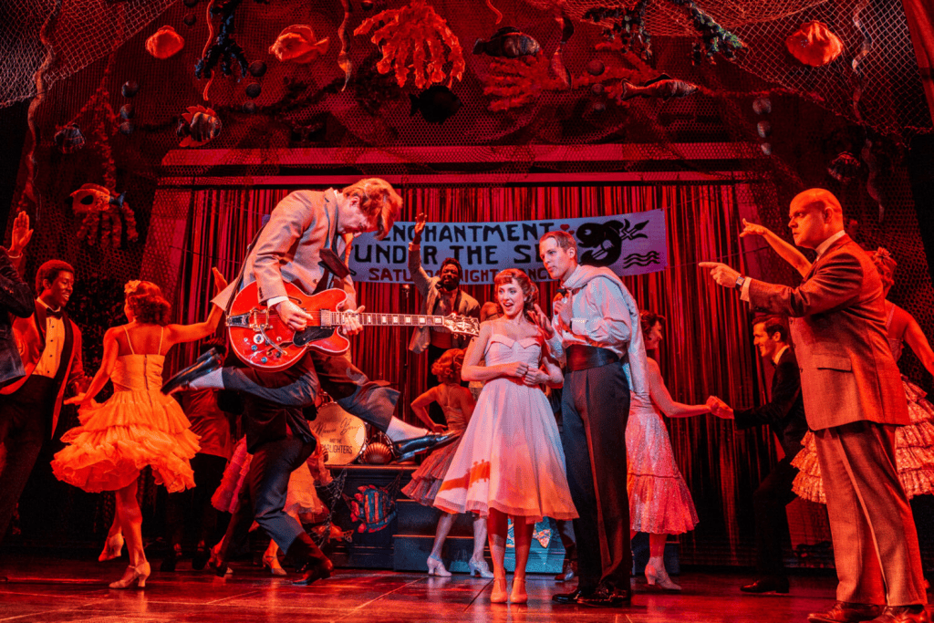 A lively stage scene from a musical performance featuring dancers in colorful costumes, a guitarist in mid-air playing a red guitar, and a backdrop with the text 'Entertainment Under The Sea'.