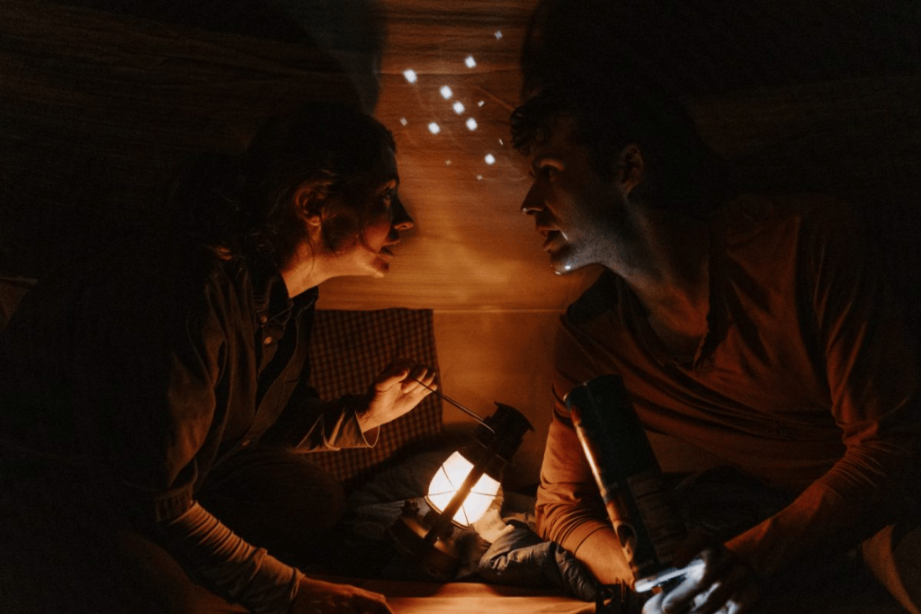 Two actors engage in an intense conversation inside a tent, illuminated by a lantern and flashlight, creating a dramatic atmosphere.