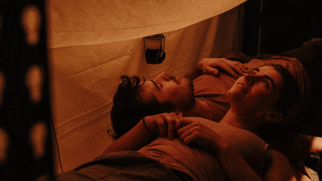 A couple lying together under a makeshift tent illuminated by a lantern, sharing a relaxed moment.