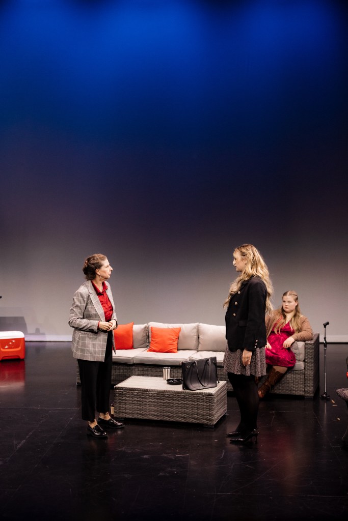 “A War Is Coming”: Voodoo’s Heroes of the Fourth Turning 2 Two actresses perform a scene on stage, one wearing a checkered blazer and red shirt, while the other is in a black coat and skirt. A third actress sits in the background, wearing a pink dress, alongside a modern sofa with orange cushions.