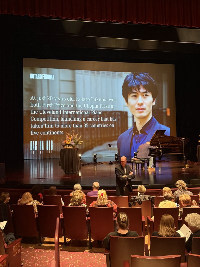 Kotaro Fukuma Opens Bachauer’s 2026–27 Season with Homage to Liszt 3 An audience seated in a theater during a piano competition event, with a large screen displaying Kotaro Fukuma's image and biography in the background. A pianist is seen performing on stage.