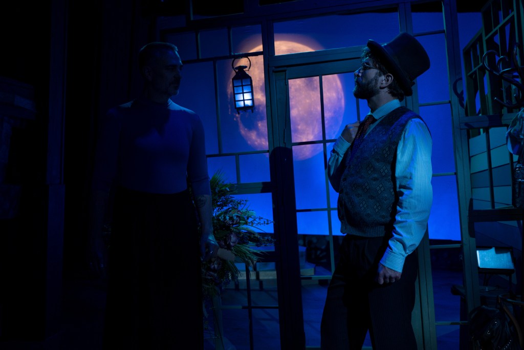 Two male actors on stage in dramatic lighting, one in a blue shirt and the other in a vintage outfit with a top hat, with a large full moon visible in the background.