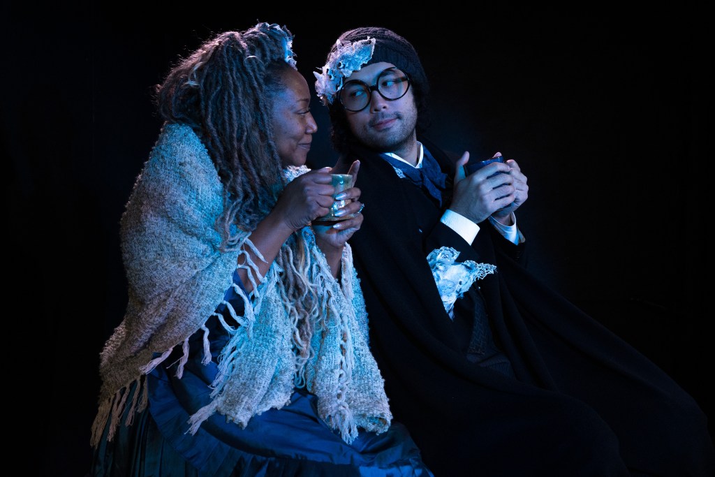 A woman and a man sitting closely together, sharing a warm moment while holding cups. The woman has long curly hair and a knitted shawl, while the man wears glasses and a knit beanie, both appearing cozy in a dimly lit setting.