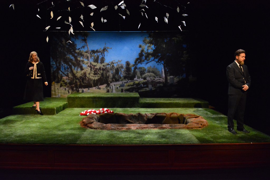 Carol Lynn Pearson's Facing East Returns, Still Asking Utah What Love Costs 6 A stage set depicting a grave site with two characters, a woman in a black dress and a man in a suit, standing next to an open grave surrounded by grass and a backdrop of a cemetery.