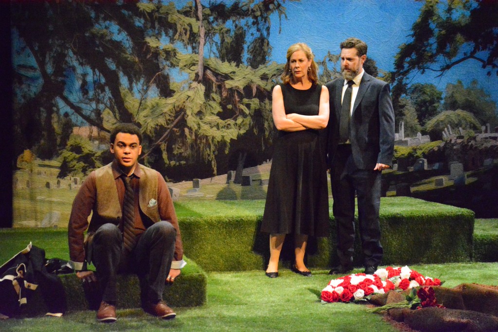 Carol Lynn Pearson's Facing East Returns, Still Asking Utah What Love Costs 3 A theatrical scene set in a cemetery with three characters: a man sitting on the ground in a brown vest and tie, a woman in a black dress standing with her arms crossed, and a man in a suit next to her. They are surrounded by grass and a pile of roses.