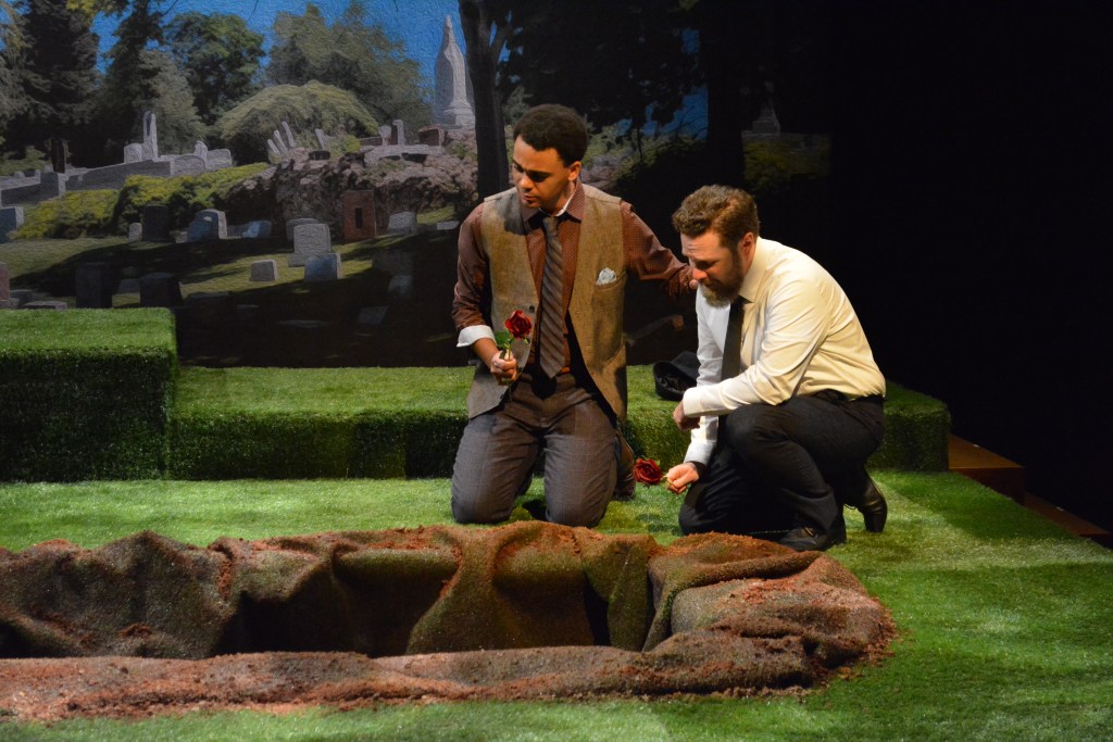 Carol Lynn Pearson's Facing East Returns, Still Asking Utah What Love Costs 5 Two men kneeling by a grave on a stage, surrounded by green grass and a backdrop of a cemetery. One man holds a rose.