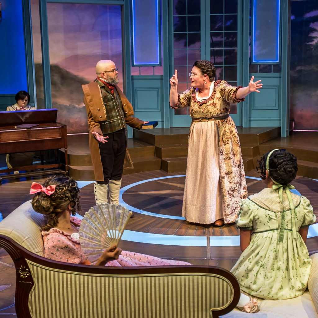 Listening to Mrs. Bennet: Larson’s Pride and Prejudice at The Ruth 3 A theatrical scene featuring a woman in a floral dress passionately gesturing while speaking to a man in period costume. In the background, a woman plays the piano, and two seated women in period attire observe the discussion.