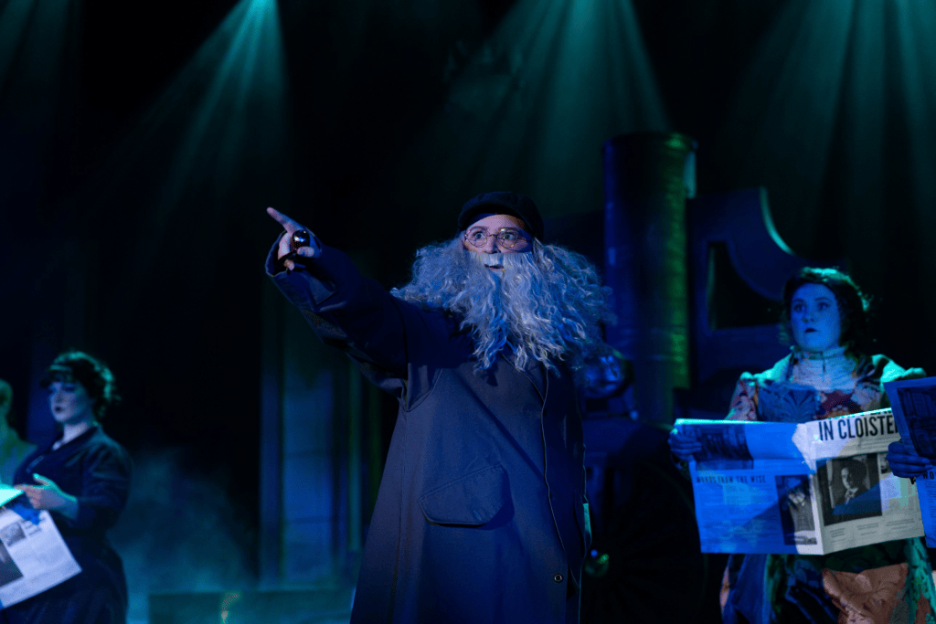 A performer wearing a long coat and glasses with a large grey beard points dramatically on stage, while others hold newspapers in a theatrical setting illuminated by blue lights.