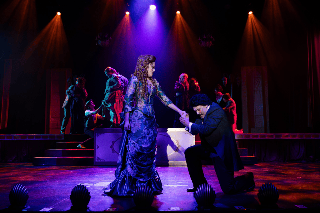 A theatrical performance featuring a woman in a blue gown and a man in a black suit, with dramatic lighting and a cast of performers in the background.