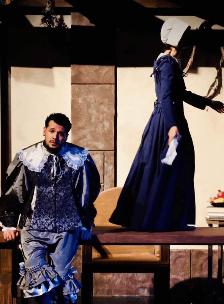 Painted in Light, Driven by a Pulse: PYGmalion's Witch by Jen Silverman 5 A man in elaborate historical costume sitting on a table while a woman in a long dress and bonnet walks away in a dimly lit interior setting.