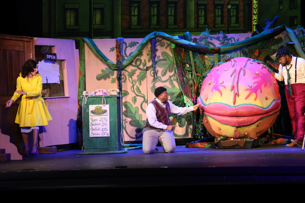 A theatrical scene featuring three characters on stage; a woman in a yellow dress stands to the left, while a man kneels in front of a large, colorful plant, and another man stands to the right, all set against a backdrop of urban buildings and decorative foliage.