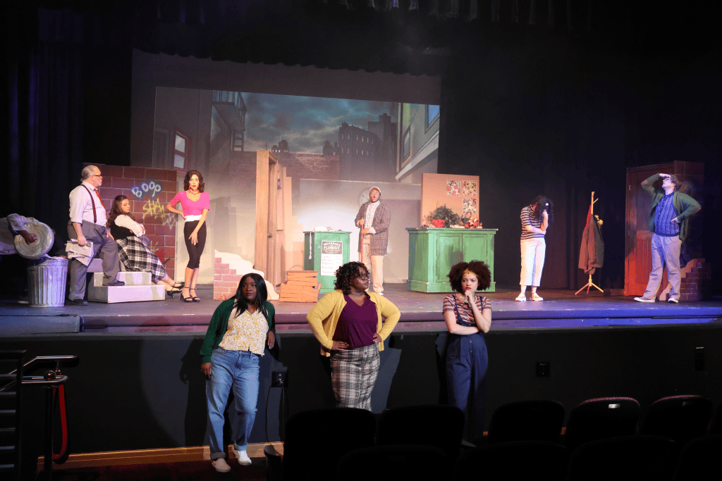A theater stage with actors performing a dramatic scene. The backdrop features a cityscape, while various props, including a trash can and colorful set pieces, are arranged on stage. The actors are dressed in diverse costumes and exhibit varied emotions.