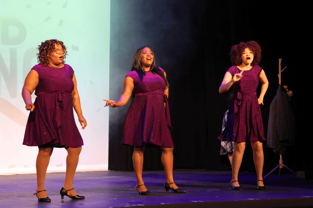 Three women performing on stage, wearing matching purple dresses, under stage lights.