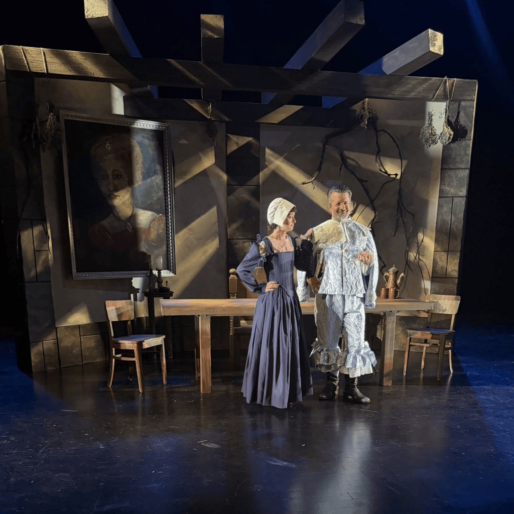 Painted in Light, Driven by a Pulse: PYGmalion's Witch by Jen Silverman 4 Two actors in period costumes pose on stage in front of a painted backdrop and wooden table, with a portrait hanging on the wall.