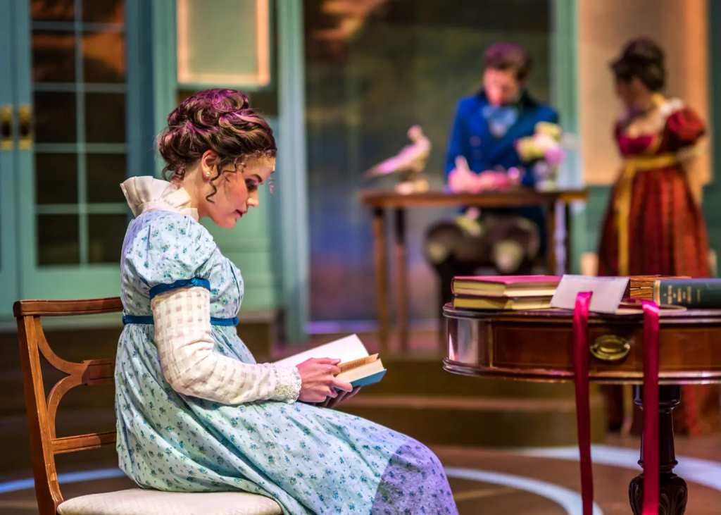 Listening to Mrs. Bennet: Larson’s Pride and Prejudice at The Ruth 6 A woman in a blue dress reads a book while two figures engage in conversation at a table in a theatrical setting.